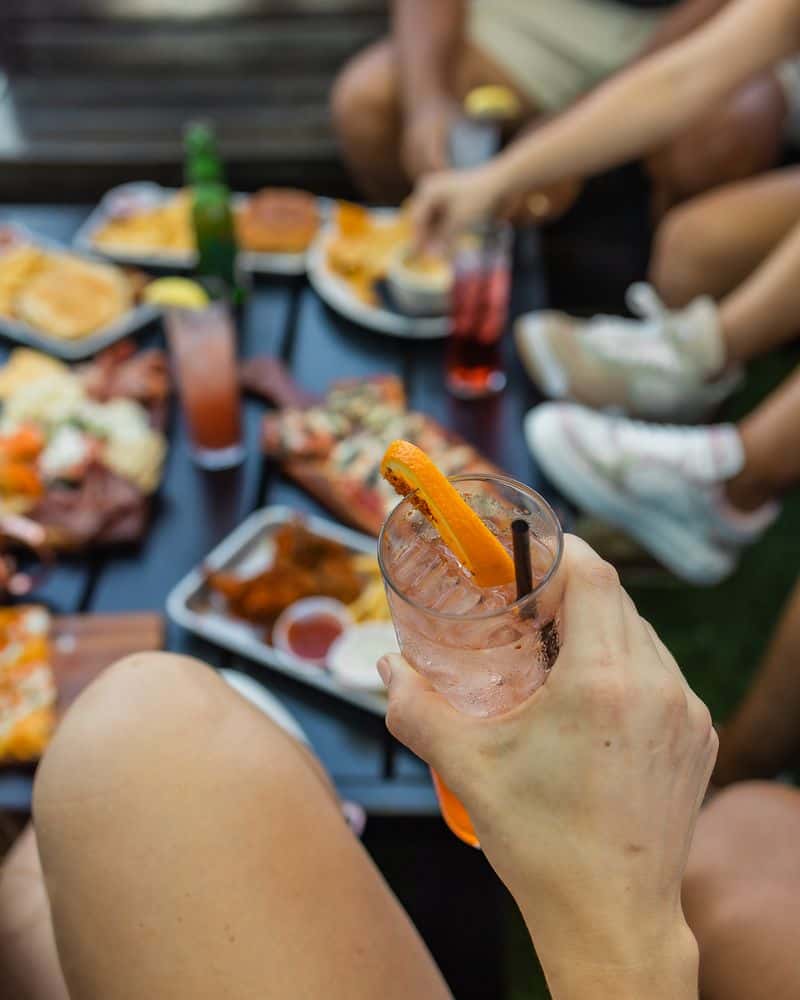 Person holding a refreshing cocktail garnished with an orange slice during an outdoor gathering, with a table full of bar food favorites like flatbread pizza, chicken wings, fries, sliders, and charcuterie in the background—perfect for a social brunch or weekend patio hangout.