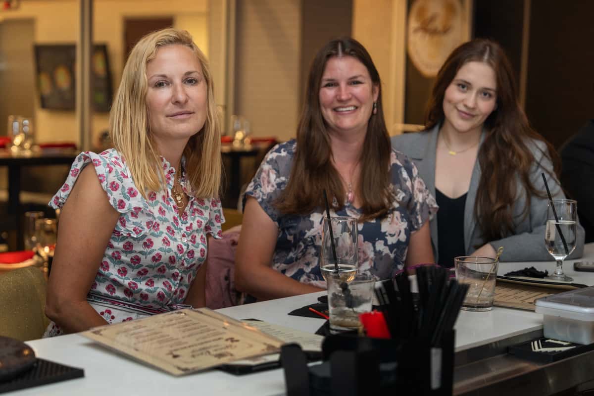 Three women with drinks and menus at a white bar counter, enjoying the vibrant atmosphere of Āne Bar & Restaurant.