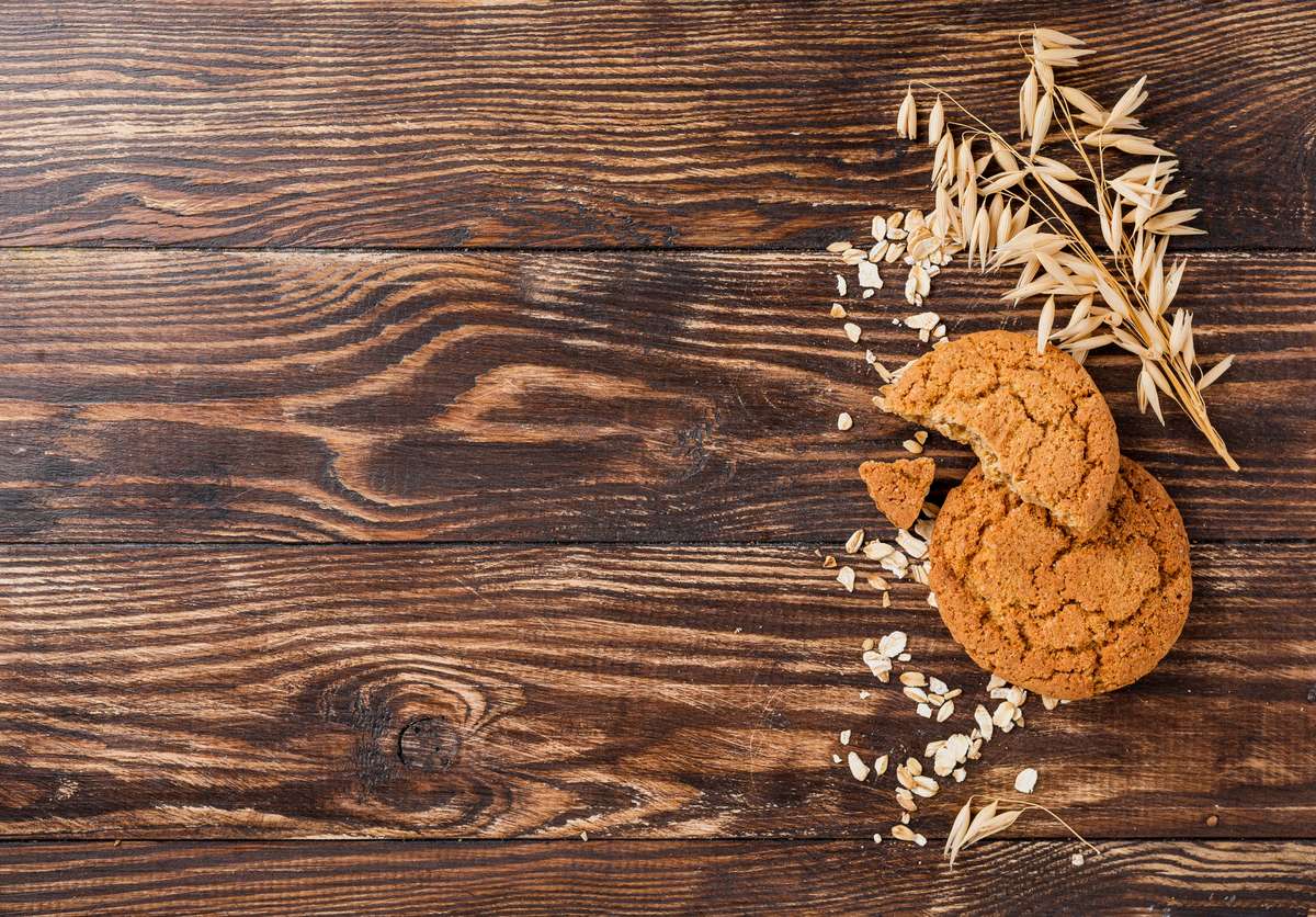 Two cookies with oat sprouts sit on a wooden table. One cookie is broken, and there are oats scattered on the table next to the cookies.