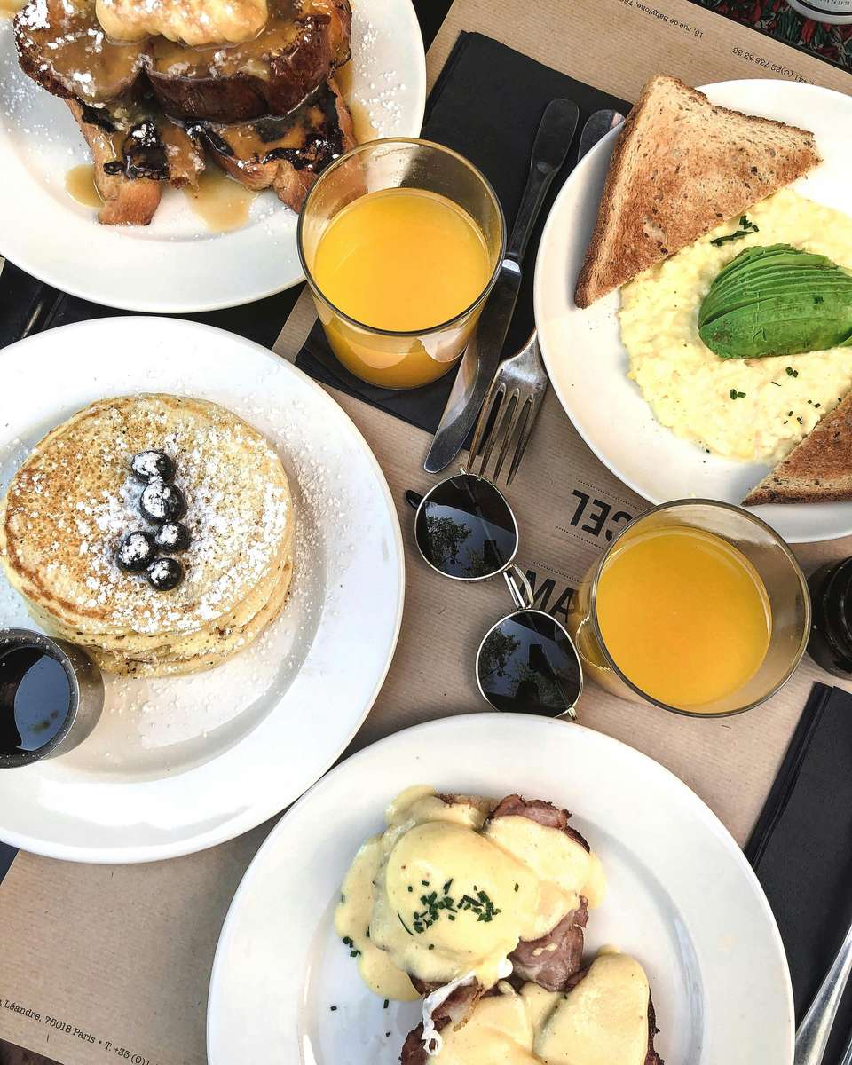 Assorted brunch dishes with pancakes, eggs benedict, avocado toast, French toast, and glasses of orange juice on a table.