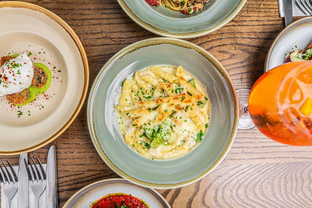Creamy baked penne pasta with herbs, surrounded by assorted gourmet dishes and a glass of orange aperitif on a wooden table.