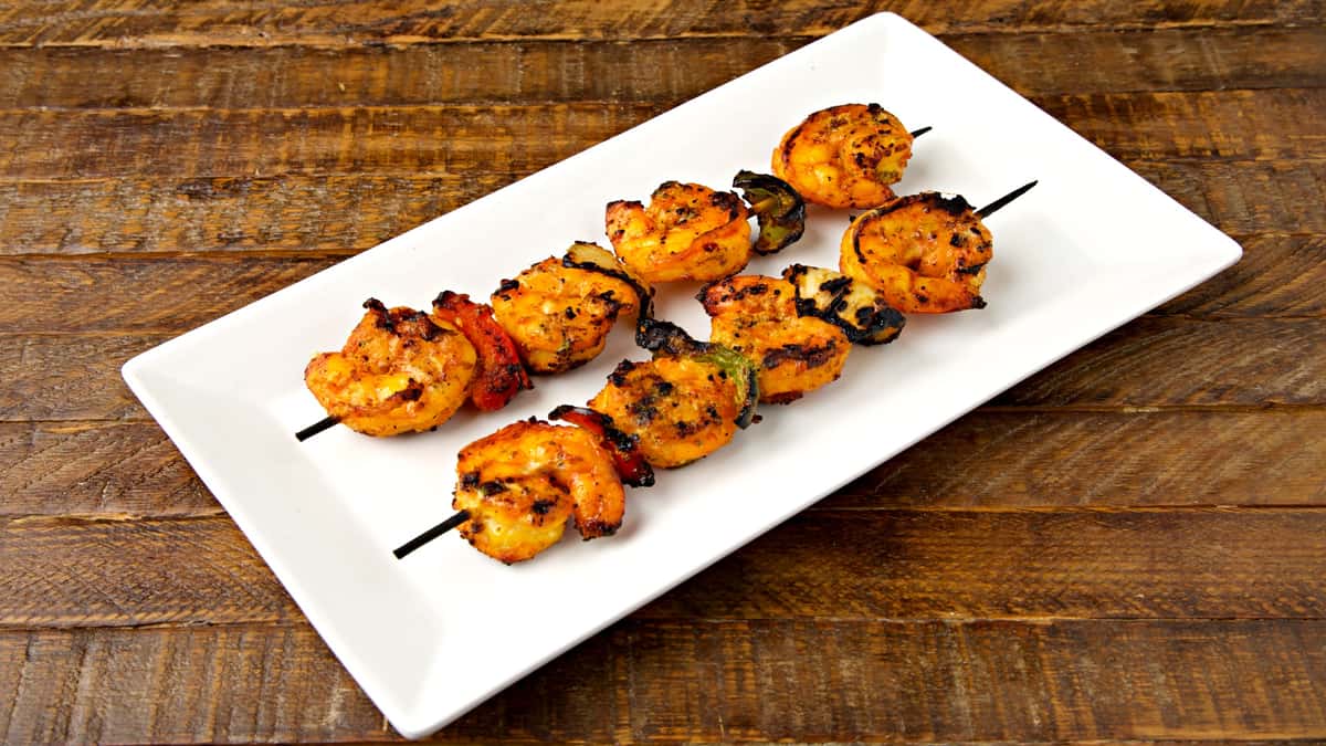 Two vibrant, char-grilled shrimp and pepper skewers served on a white rectangular platter over a rustic wooden table.