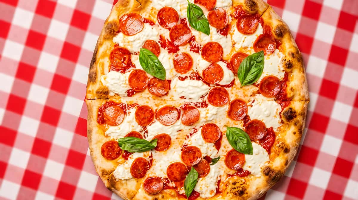 Pepperoni pizza with basil and melted mozzarella on a red and white checkered tablecloth.