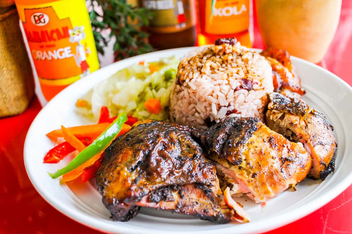A plate of Jamaican food featuring jerk chicken, seasoned rice, and a side of steamed vegetables, garnished with fresh tomatoes and green peppers.
