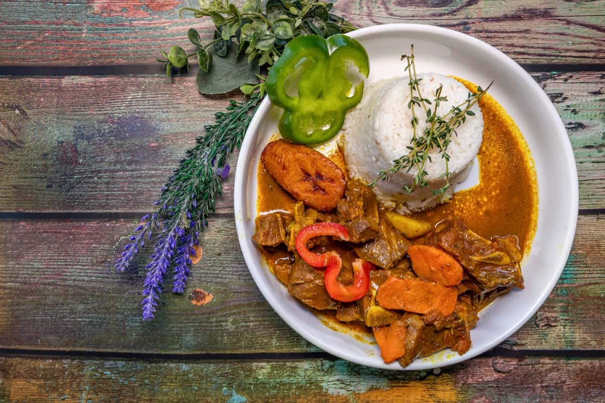Caribbean-style oxtail stew with carrots and peppers, served with white rice, fried plantain, and fresh thyme garnish.
