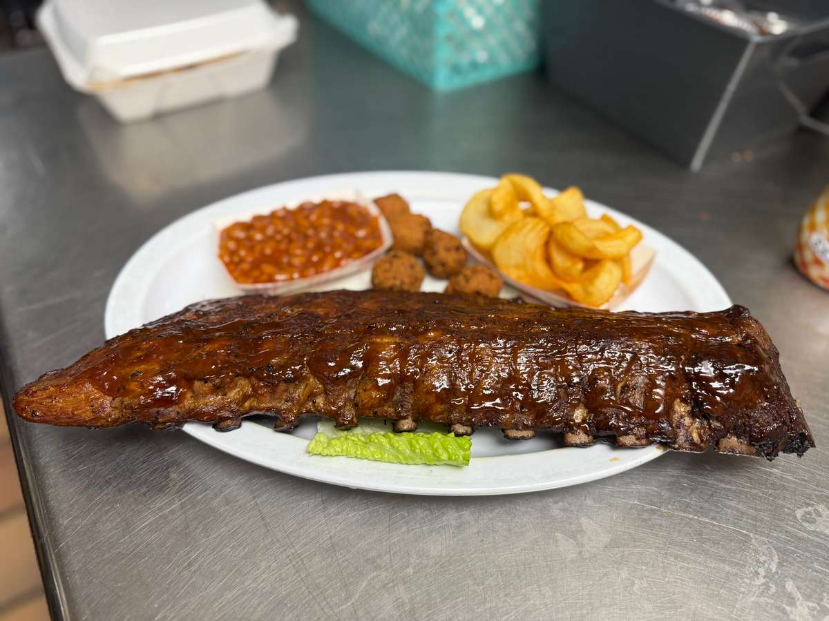 Full rack of glazed barbecue ribs served with seasoned potato chips, baked beans, and fried hushpuppies, plated for a hearty Southern-style meal.