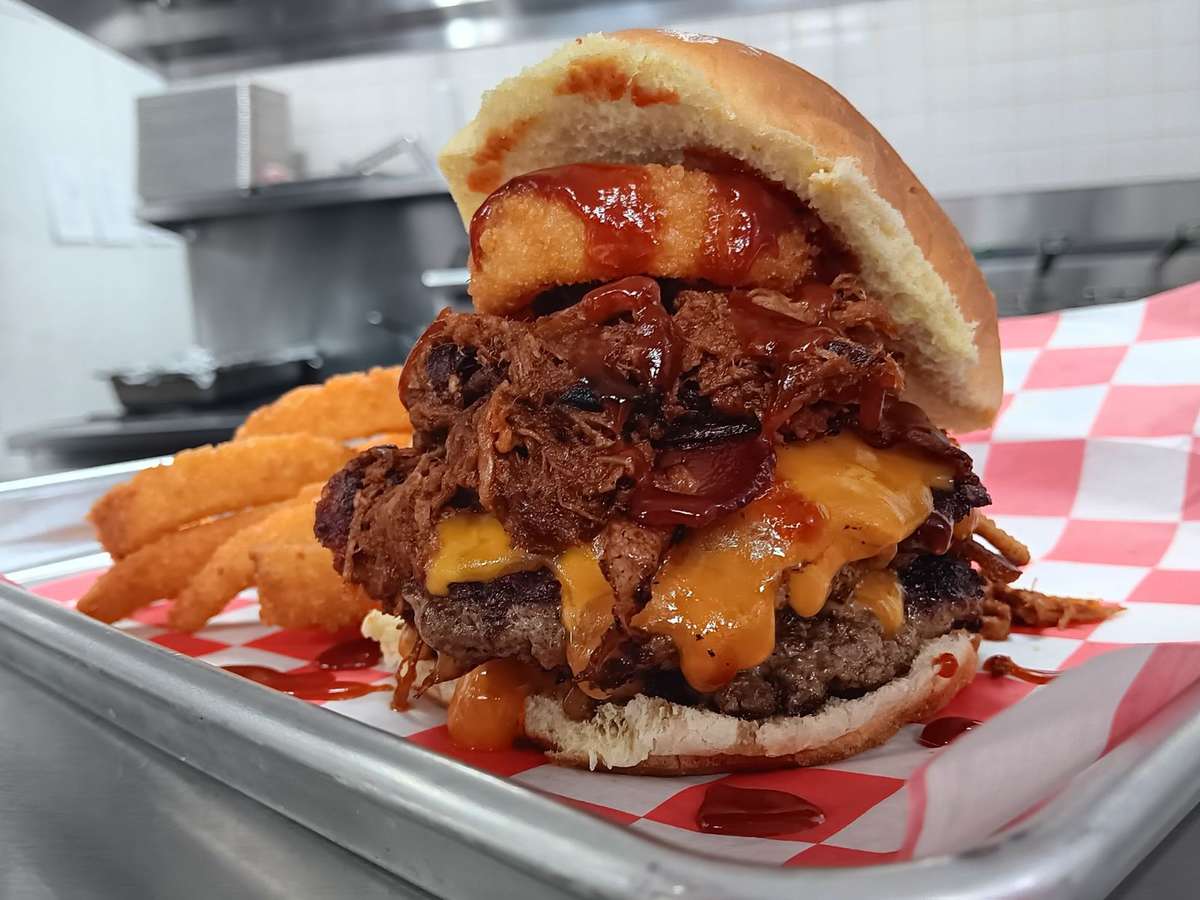 Close-up of a stacked barbecue cheeseburger with melted cheddar, slow-cooked pulled pork, crispy onion rings, and tangy barbecue sauce on a toasted bun, served with golden fried onion rings on a red checkered tray in a diner-style kitchen.