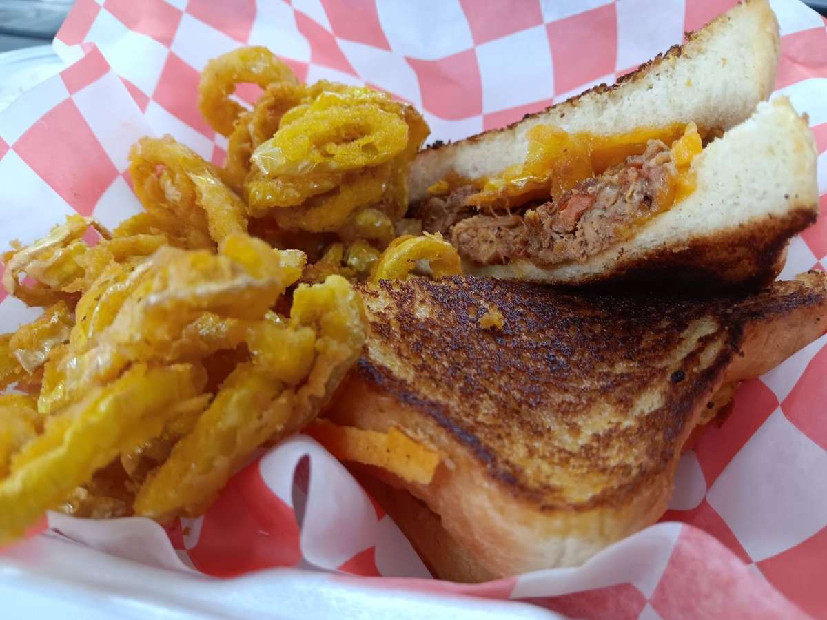 Toasted pulled pork grilled cheese sandwich with melted cheddar on white bread, served with a side of crispy golden onion straws in a red checkered basket.