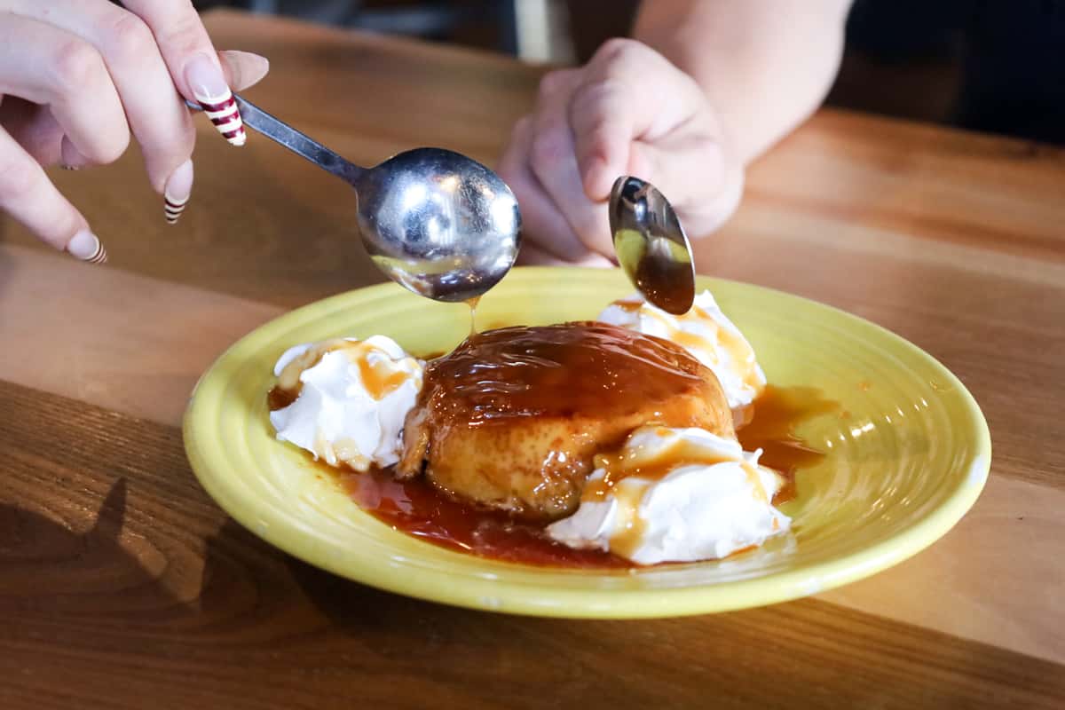 A golden flan dessert topped with caramel sauce and whipped cream on a bright yellow plate, with two people about to share it with silver spoons.