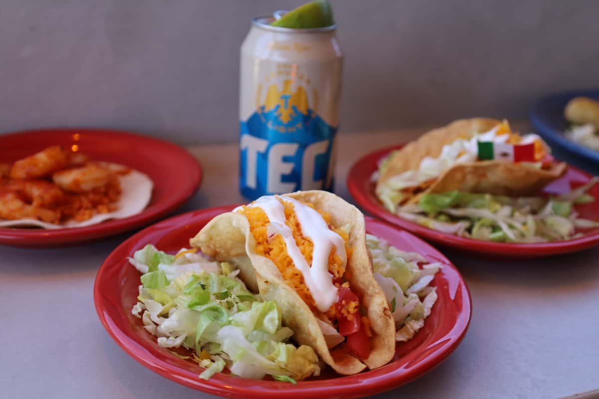 Three red plates of Mexican tacos topped with shredded cheese and sour cream, served alongside a Tecate Light beer.