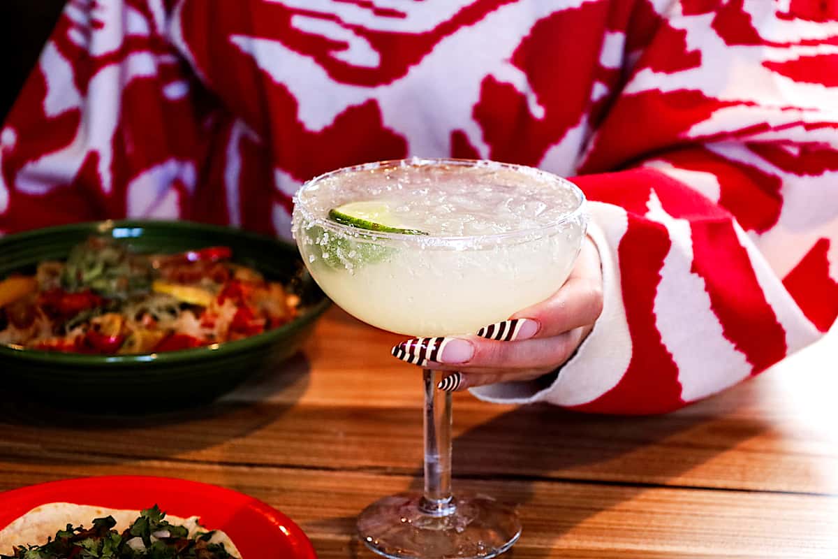 A person in a red and white patterned sweater holds a lime margarita with a salt rim at a wooden table served with Mexican dishes.