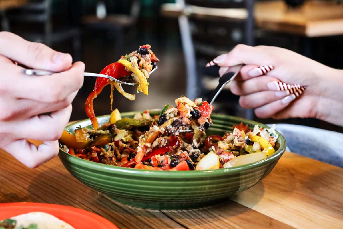 Two people share a vibrant Mexican-style bowl filled with red peppers, yellow peppers, and black beans from a green ceramic dish on a wooden table.