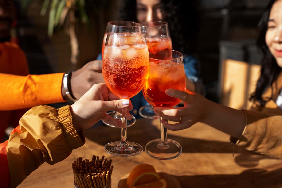 A close-up of several people toasting with large glasses of a refreshing orange cocktail, filled with ice and garnished with a slice of orange.