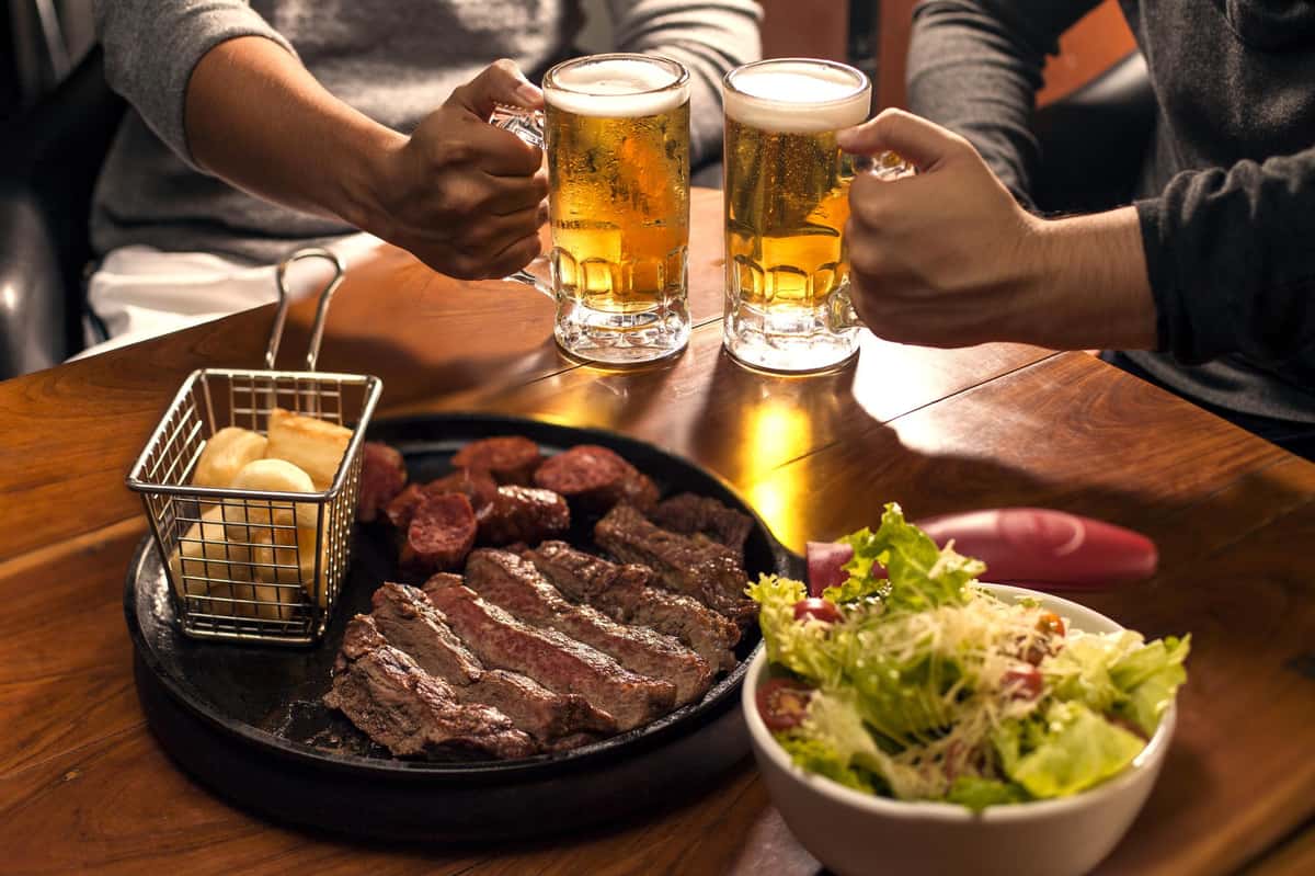 wo people toast with beer, while enjoying grilled steak and accompanied by a basket of bread rolls and a fresh salad.