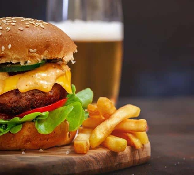 Cheese burger with lettuce, tomato, spicy sauce and jalapenos on a seeded bun with French fries and a beer in the background