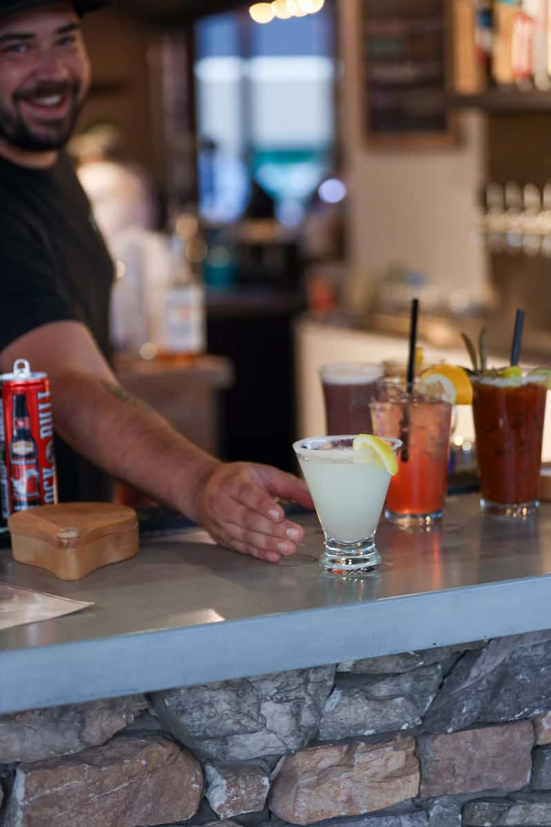 Smiling bartender at Otto's Landing serving cocktails on the stone bar counter, including a margarita with lemon, a Bloody Mary, and a refreshing citrus drink with orange garnish.