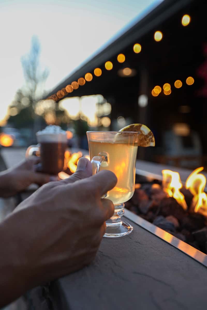 Close-up of a warm cocktail with a lemon garnish held near a glowing outdoor fire pit at Otto's Landing, with string lights and a cozy patio ambiance at sunset—ideal for evening drinks and casual outdoor dining.