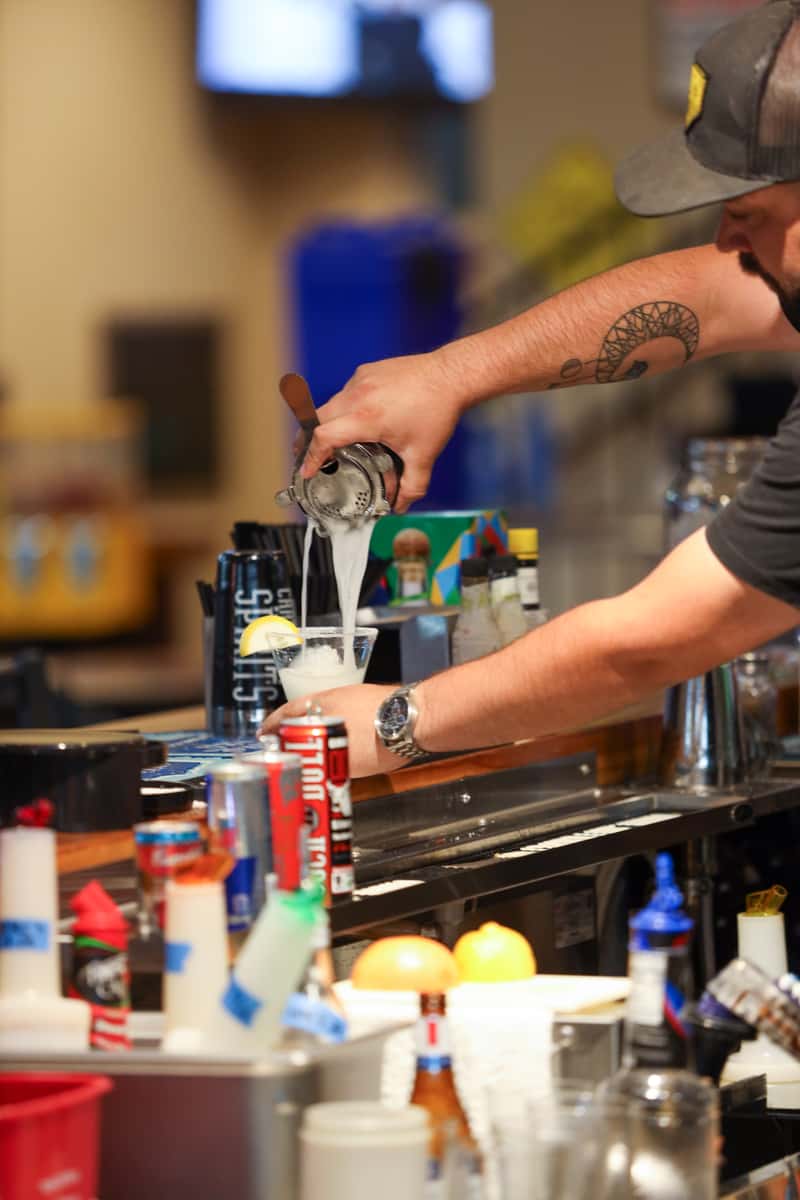 Bartender pouring a fresh cocktail at Otto's Landing bar, surrounded by bottles and garnishes, showcasing handcrafted drink service.
