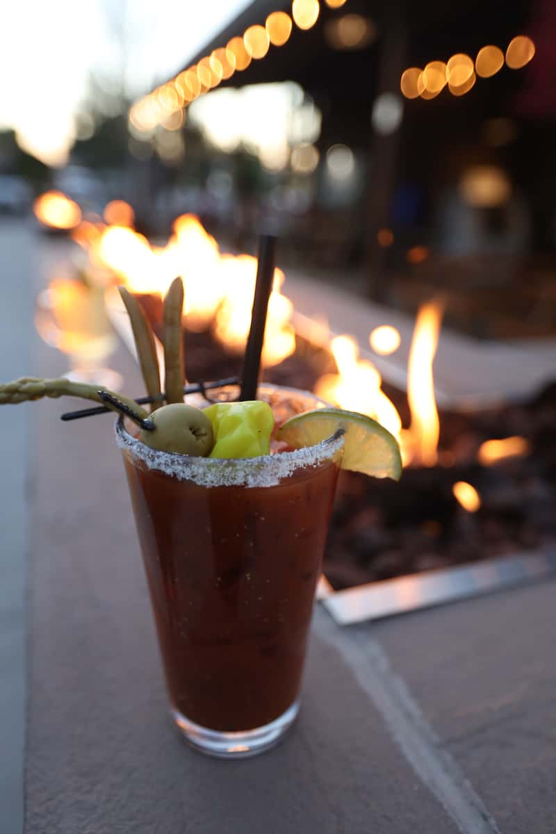 Bloody Mary cocktail with olives, lime, and pickled vegetables resting on the edge of Otto's Landing's fire pit patio at sunset.