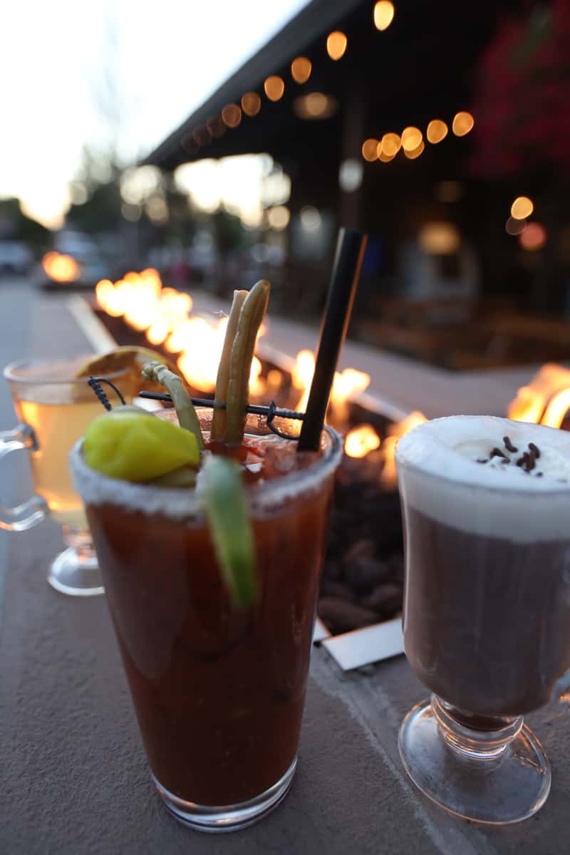 Bloody Mary cocktail and whipped-cream-topped hot cocoa placed beside Otto's Landing's outdoor fire pit during golden hour.