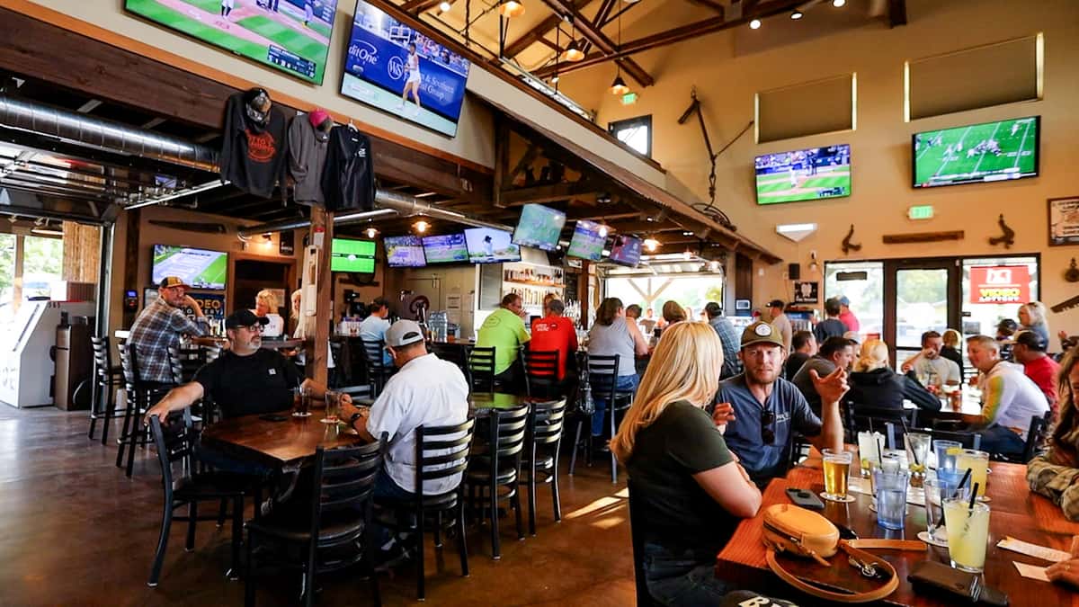 Guests enjoying drinks and conversation inside Otto's Landing sports bar, with multiple flat-screen TVs showing live games, rustic wood accents, and a lively crowd around the bar and tables.