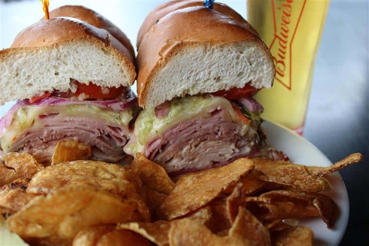 large sandwich with turkey, roast beef and ham on a roll with potato chips and a glass of beer