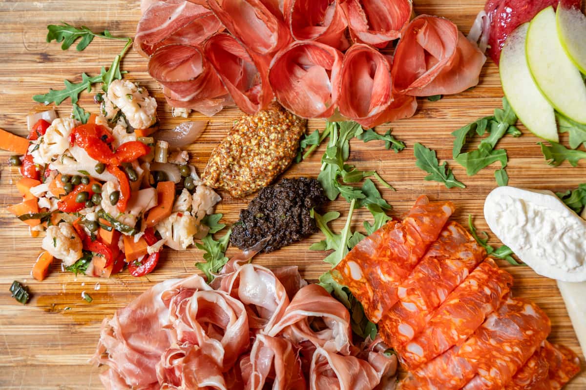 Overhead view of a gourmet Italian charcuterie board with sliced prosciutto, spicy salami, coppa, artisan cheeses, fresh apple slices, giardiniera, grainy mustard, black olive tapenade, and arugula on a rustic wooden board—perfect for antipasto platters or wine pairings.