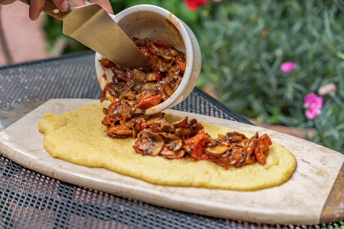 Hand spooning a hearty mushroom and tomato ragu onto a bed of creamy polenta, served on a rustic stone board outdoors—perfect for showcasing traditional Northern Italian comfort food or al fresco dining experiences.