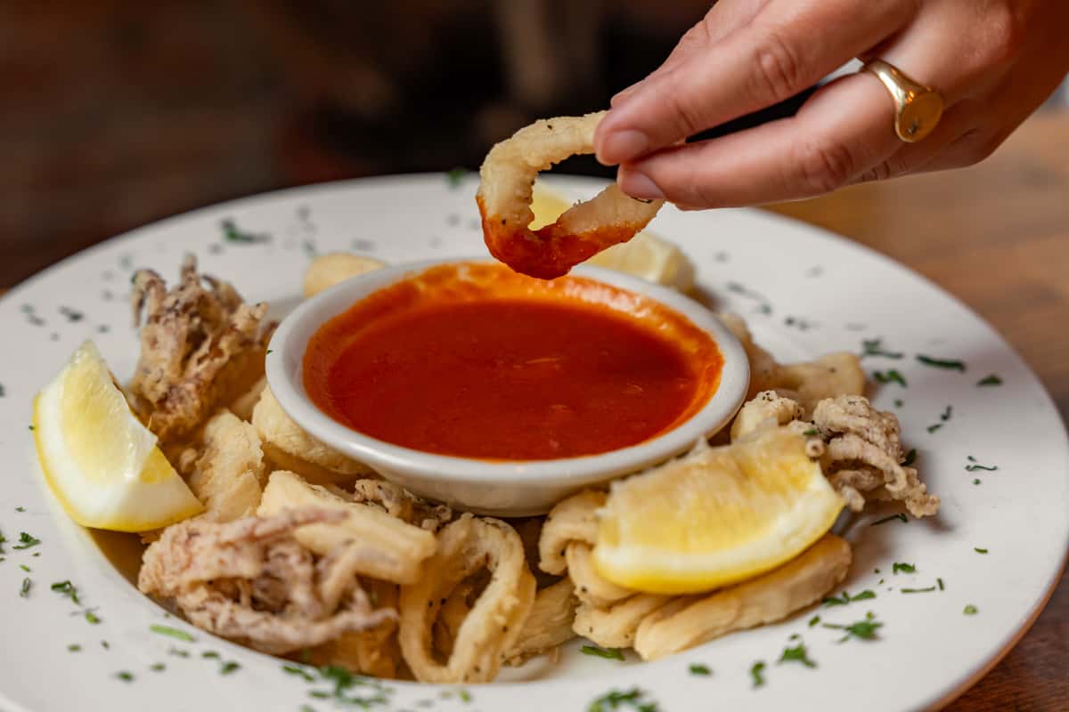 Close-up of a hand dipping crispy fried calamari into zesty marinara sauce, served with lemon wedges on a white plate garnished with herbs—classic Italian appetizer perfect for sharing.
