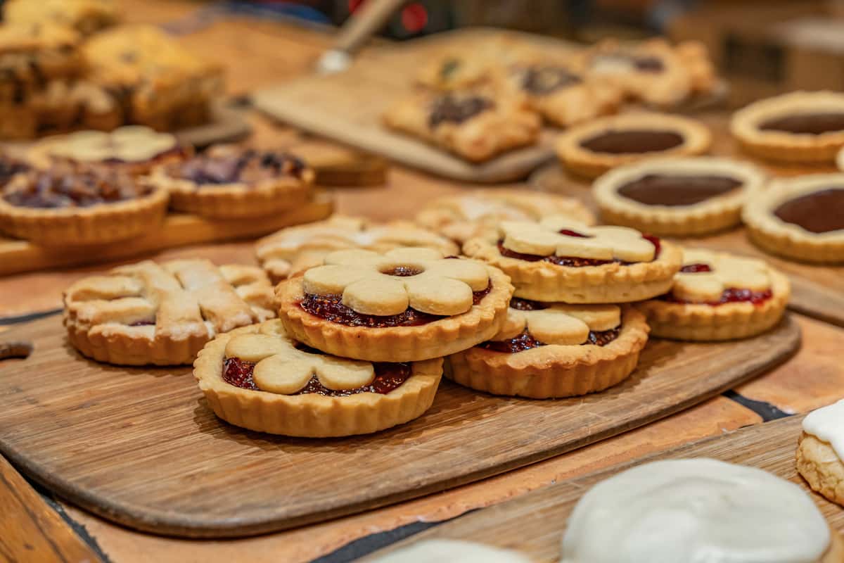 Assortment of buttery jam-filled cookies topped with daisy-shaped shortbread, arranged on a rustic wooden tray—ideal for a bakery display or seasonal dessert menu.
