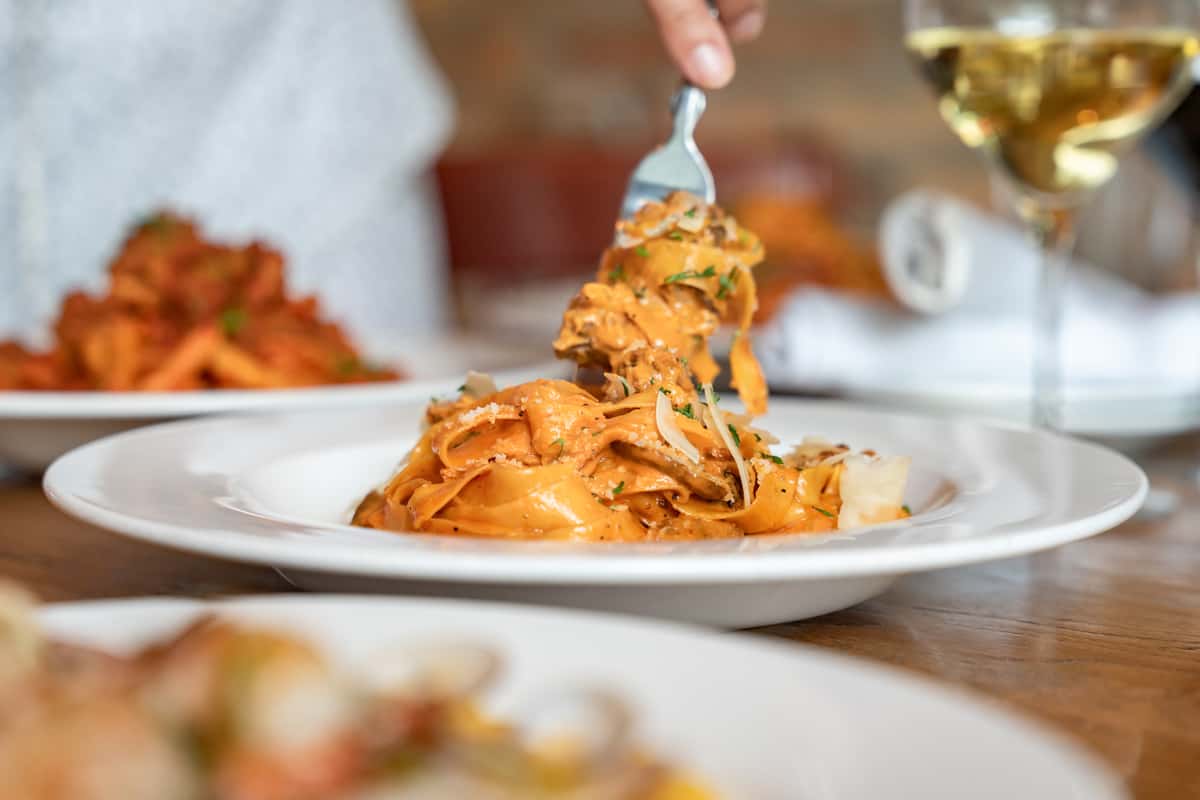Close-up of creamy tagliatelle pasta being twirled on a fork, served with shaved Parmesan and fresh herbs, with a glass of white wine and additional pasta dishes blurred in the background—perfect for an elegant Italian dining experience.