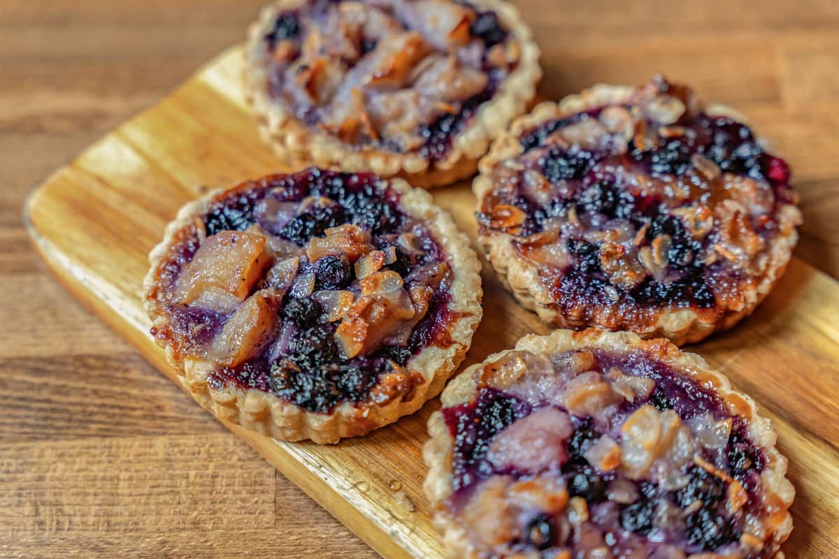 Close-up of rustic mini tarts filled with a baked fruit medley, including dark berries and diced pears, on a golden flaky crust—ideal for a bakery display or seasonal dessert offering.