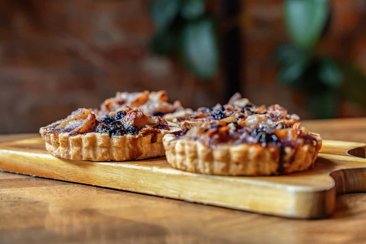 Side view of artisanal fruit tarts topped with a glossy blend of dark berries and diced pear, displayed on a wooden serving board against a rustic backdrop.
