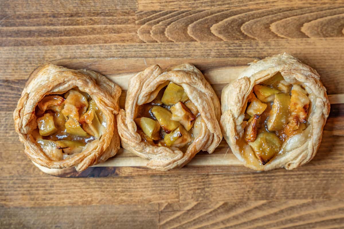 Top-down view of golden-baked apple puff pastries with caramelized filling, nestled in flaky, swirled pastry shells on a wooden board—perfect for fall or farmhouse-style dessert menus.