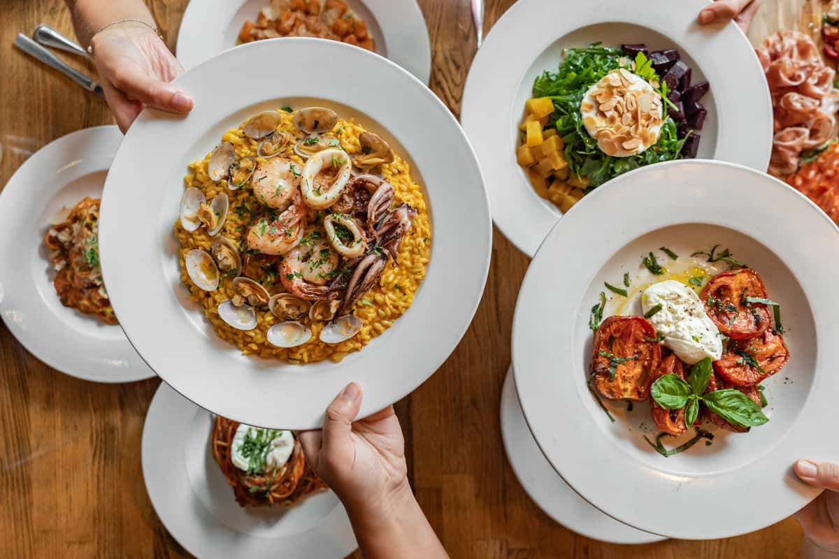 Top-down view of a vibrant Italian seafood risotto plated with shrimp, clams, and calamari, surrounded by fresh Mediterranean dishes including burrata with grilled tomatoes, a beet and mango salad, and pasta entrées, all served on white plates over a wooden table.
