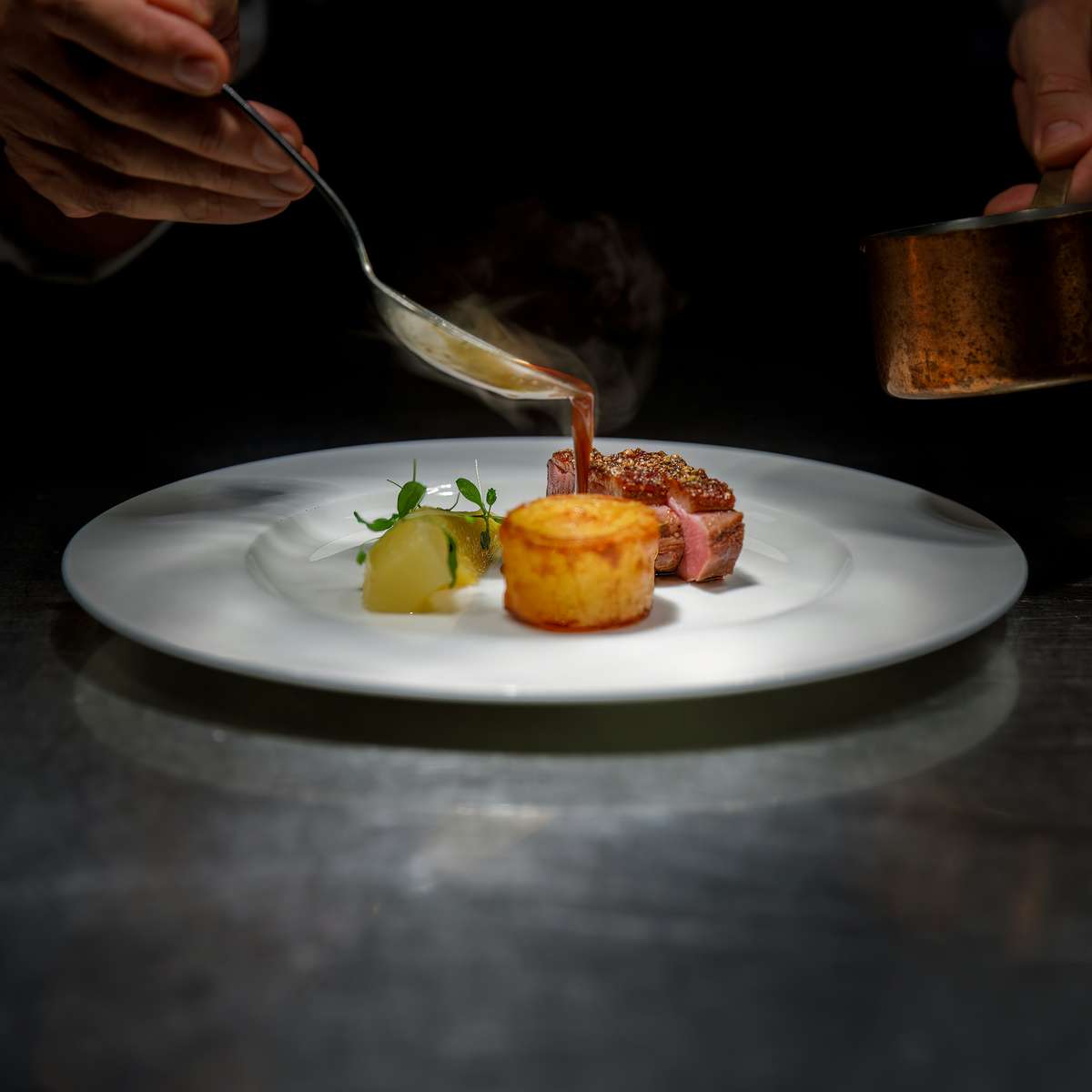 Gourmet dish being plated by a chef under spotlight, featuring seared meat, a golden potato cake, and elegant garnishes on a white plate in a high-end restaurant.