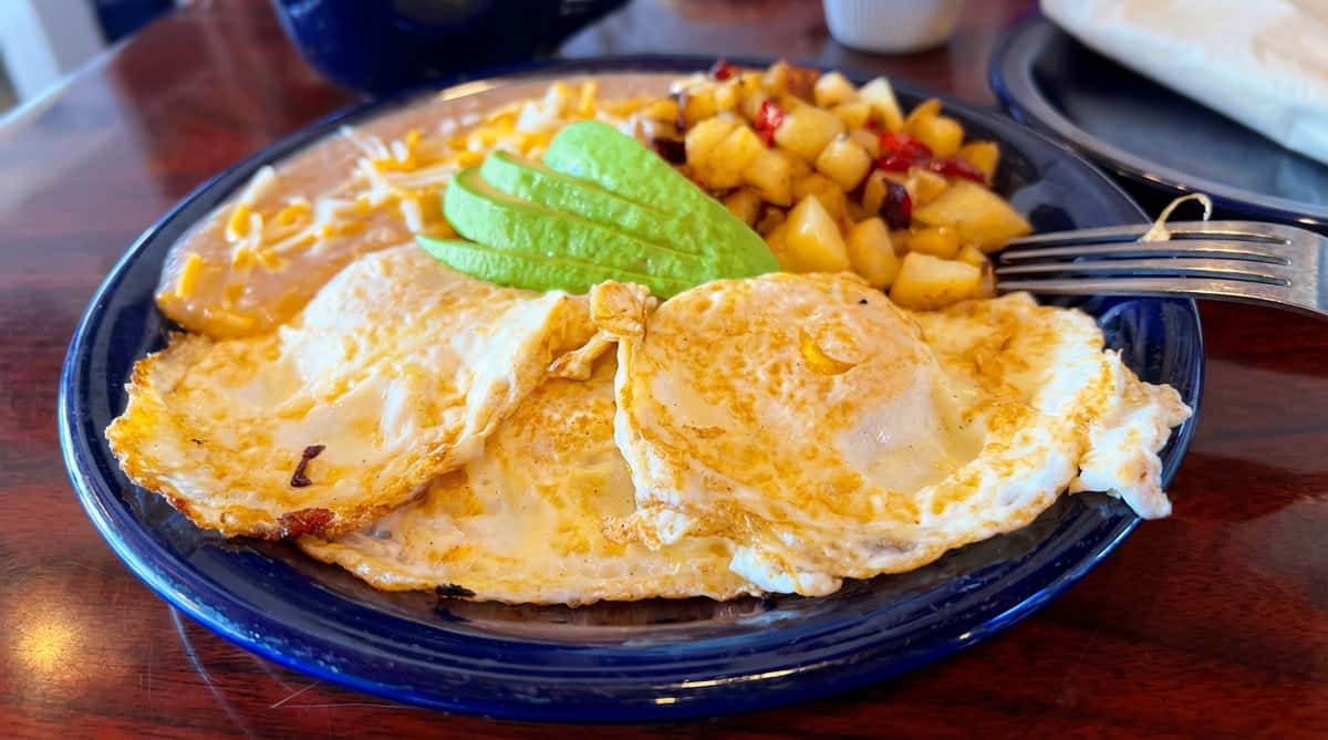 Breakfast plate with fried eggs, refried beans with cheese, avocado slices, and home fries.