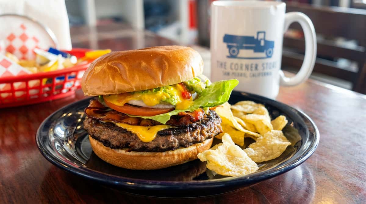Burger with melted cheese, bacon, lettuce, tomato, onion, and guacamole, served with potato chips and a mug.