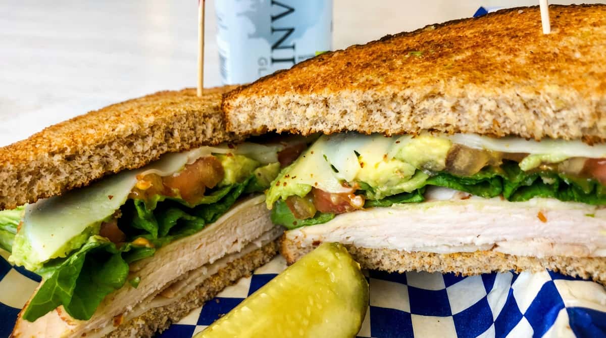 Turkeyclub sandwich on toasted wheat bread with lettuce, tomato, avocado, and cheese, served with a pickle spear.
