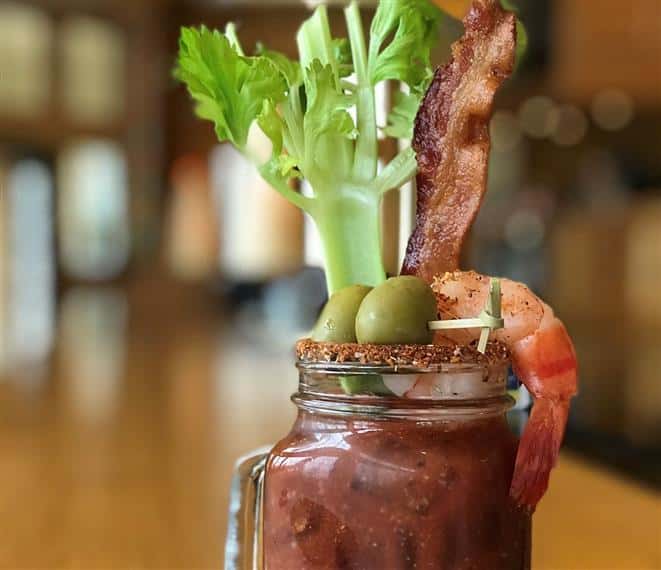 bloody mary in a mason jar with a handle, celery, shrimp, olives and bacon sticking out the top