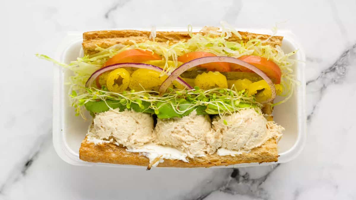 Fresh tuna salad sandwich on toasted bread, topped with shredded lettuce, tomato slices, red onion, banana peppers, sprouts, and creamy avocado slices, served in a takeout container.