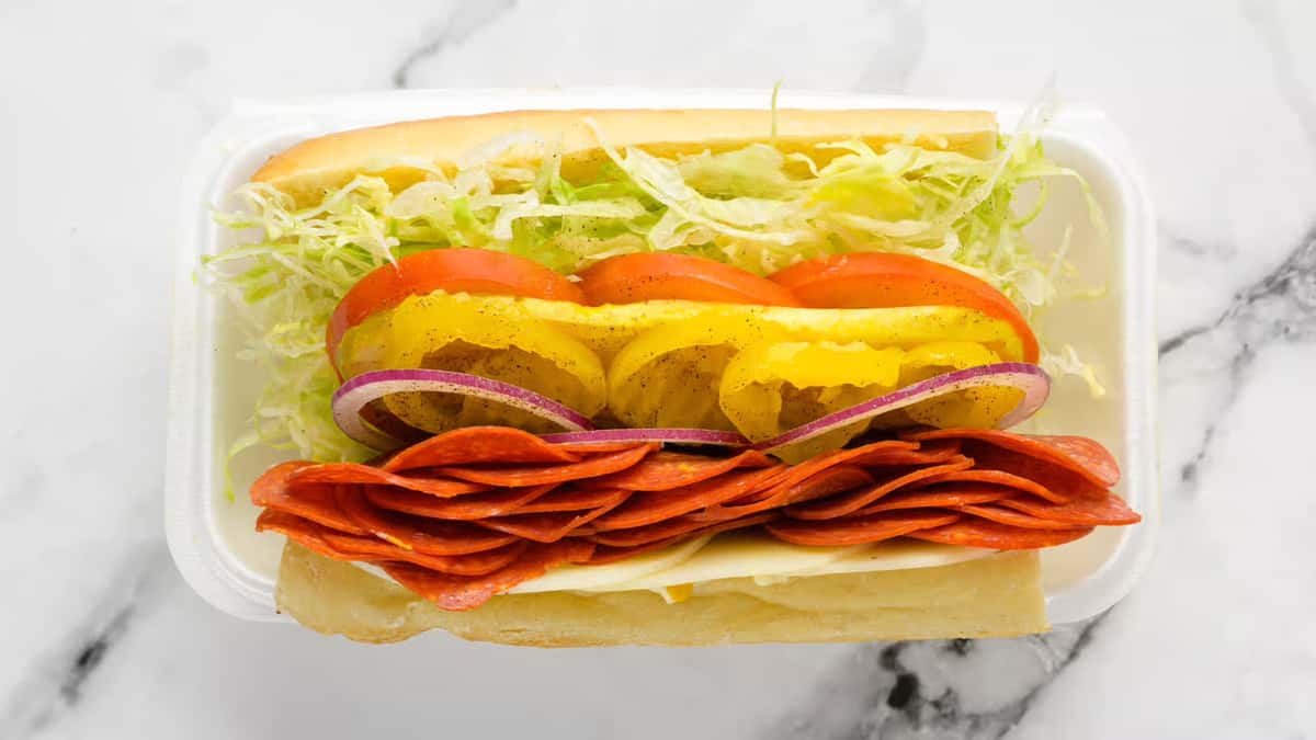 Assorted gourmet sandwiches from Eagle Sandwich Co., featuring layers of fresh deli meats, melted cheese, lettuce, tomato, pickles, and sprouts, served on toasted sub rolls with sliced cucumbers and tomatoes on the side.