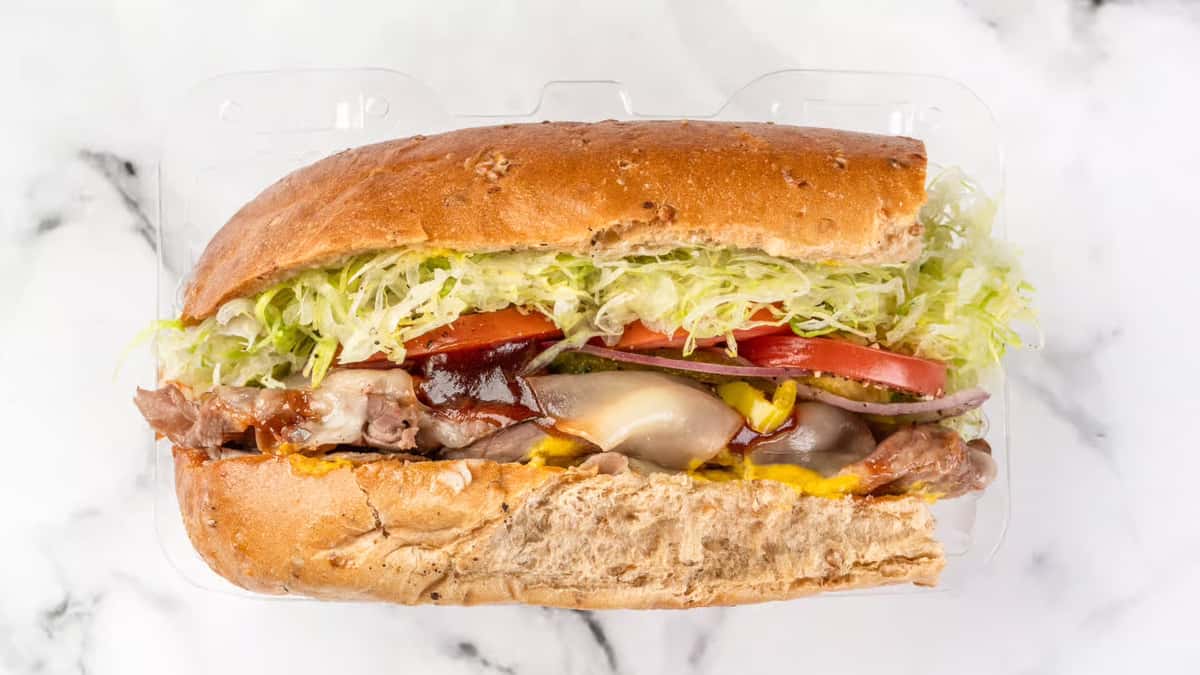 Hearty roast beef sub sandwich with melted cheese, tangy barbecue sauce, shredded lettuce, tomato slices, and red onion on a toasted wheat hoagie roll, served in a clear takeout container.