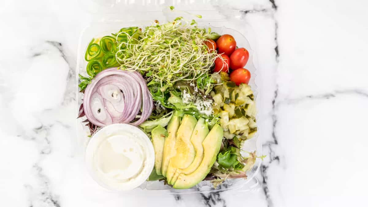 Fresh deli salad with mixed greens, avocado slices, grape tomatoes, sprouts, sliced red onions, diced pickles, and a creamy dressing on the side, served in a clear takeout container.