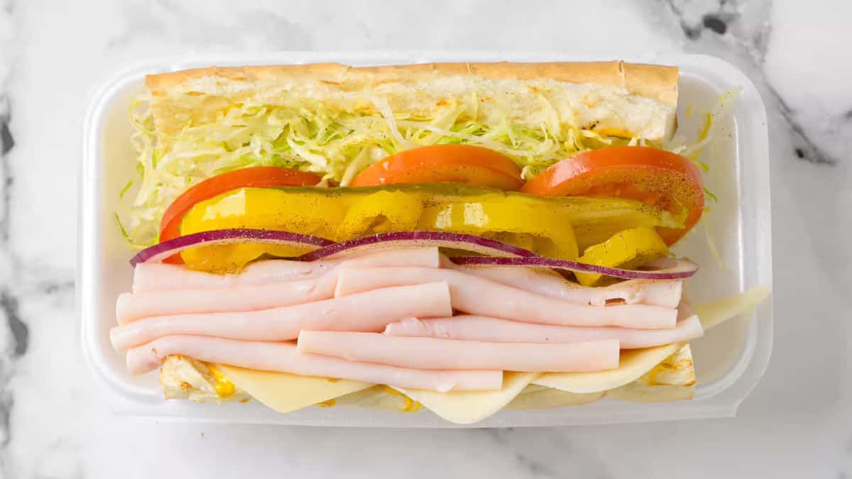 Fresh turkey sub sandwich layered with sliced turkey breast, provolone cheese, crisp lettuce, tomato, red onion, and banana peppers on a toasted hoagie roll, served in a takeout container.