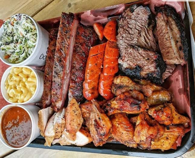 A tray filled with various meats such as chicken, ribs, and beef accompanied by macaroni and cheese, baked beans, and coleslaw 