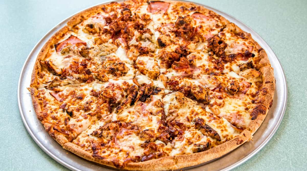 Baked pizza with melted cheese, ham, chicken, and bacon bits on a round metal tray.