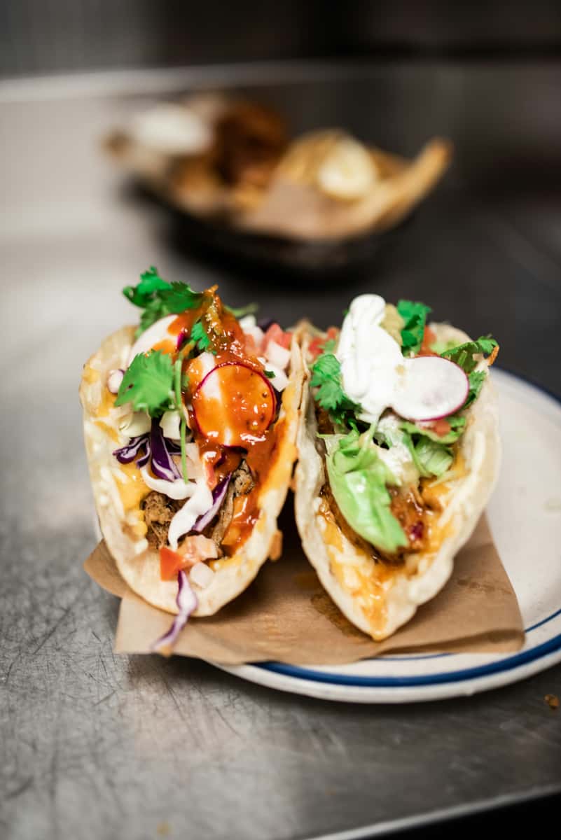 Gourmet street-style tacos filled with shredded beef, crisp lettuce, radish slices, purple cabbage, pico de gallo, and topped with creamy sour cream and tangy red salsa, served on warm flour tortillas. Presented on a rustic plate with a casual kitchen backdrop, capturing an authentic Mexican taqueria vibe.