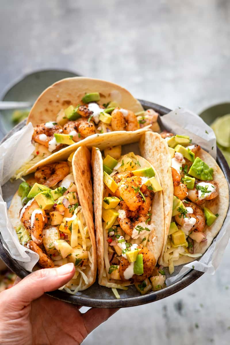 Plate of grilled shrimp tacos topped with diced avocado, shredded cabbage, pineapple salsa, and a drizzle of creamy white sauce, all served in soft flour tortillas. The vibrant seafood dish captures fresh coastal Mexican flavors in a bright, modern presentation.