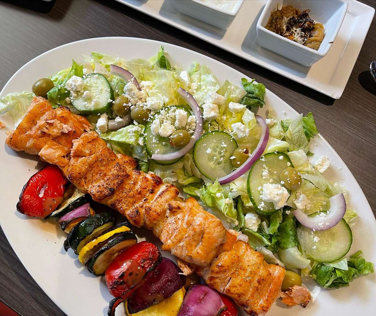 Grilled salmon and vegetable skewer served with a fresh Greek salad topped with feta, olives, cucumbers, and red onions.
